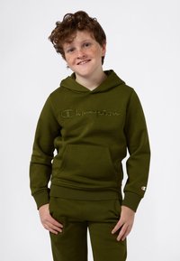 Bambino sorridente con capelli ricci e lentiggini, che indossa un felpa Champion verde e pantaloni della tuta coordinati, in piedi con le mani rilassate.