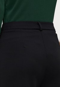 Zwarte fitted broek met een gladde textuur, voorzien van een smalle tailleband en een subtiele naaddetails aan de achterkant. Gecombineerd met een groene top.