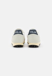 Vit sneaker med beige mockadetaljer, kraftig gummisula och marinblå häldtillbehör med "VEJA"-logotyp. Texturerad ovandel med meshavsnitt.