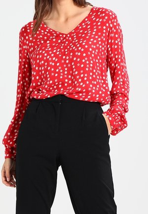Blouse rouge avec motif d'étoiles blanches, manches longues et col en V. Associée à un pantalon noir taille haute, avec des poches latérales.