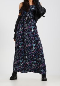 Robe longue en tissu noir avec imprimé floral coloré. Présente un design épaules dénudées, une taille cintrée avec ceinture, et un ourlet à la cheville.