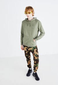 Felpa verde con tasca frontale e cordino, abbinata a pantaloni cargo mimetici marrone e verde. Sneakers nere e occhiali da sole gialli completano il look.