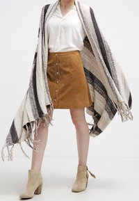 Mujer vestida con una blusa blanca, una falda de ante marrón con botones en la parte delantera, botas de tobillo beige y un gran chal de punto a rayas con bordes desflecados.