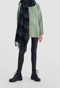 Groene kabelgebreide trui, oversized, gecombineerd met een zwart-groene geruite sjaal, zwarte neplederen leggings en zwarte veterschoenen tot de enkel.