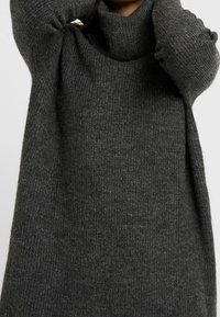 Maglione oversized grigio scuro con collo alto, texture a coste e maniche lunghe. Il materiale sembra morbido e accogliente, adatto per il clima più freddo.