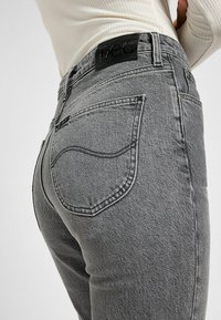 Jeans de mezclilla gris con tejido texturizado, cintura alta, pierna ajustada y costuras en contraste. Presenta un parche de cuero negro con el logotipo en la parte posterior.