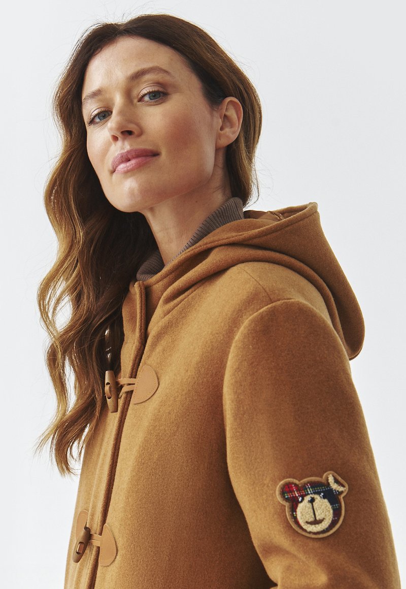 Brązowy wełniany płaszcz z kapturem, z zapięciem na klipsy, żebrowanym kołnierzem i naszywką w kształcie niedźwiedzia na prawym rękawie. Miękka tekstura i ciepły design.