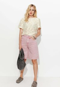 Haut crème à motifs en dentelle, shorts en denim rose et sac à main gris. Des chaussures à enfiler au motif léopard complètent la tenue. Fond lumineux.