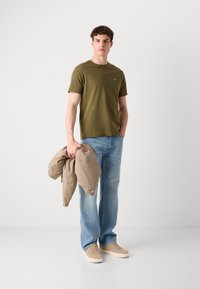 T-shirt vert olive à manches courtes, jeans en denim bleu clair, veste beige tenue à la main, baskets beiges, se tenant devant un fond neutre.