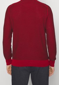 Pull rouge en maille avec un motif texturé, poignets et ourlet côtelés, et détail de surpiqûres contrastantes sur les côtés. Porté avec un pantalon gris foncé.