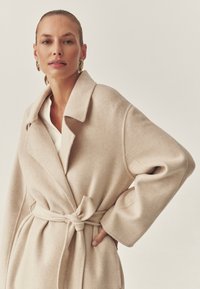 Bež volnen plašč z ovijanjem, povezovan s pasom, veliki zavihani ovratnik in široki rokavi. Mehka tekstura in sproščen kroj, brez odvečnih detajlov.