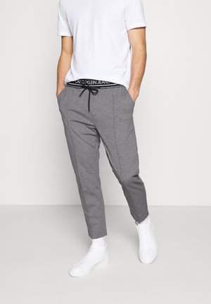 Mężczyzna w białej koszulce, szarych spodniach ze sznurkiem i widocznym paskiem Calvin Klein Jeans oraz białych trampkach stoi z rękami w kieszeniach.