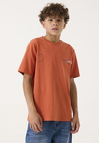 Krustfarbendes Baumwoll-T-Shirt mit kurzen Ärmeln, Rundhalsausschnitt und bedruckten Akzenten auf der linken Brust und dem Ärmel. Darunter sichtbare Denim-Shorts.