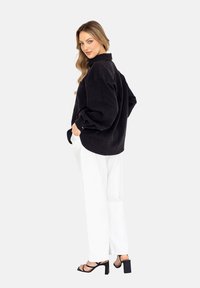 Czarna koszula z korduroy w oversize'owym fasonie z kołnierzykiem i listwą guzikową, zestawiona z białymi spodniami o szerokich nogawkach i czarnymi sandałami na obcasie.