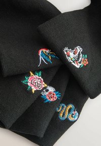 Vier schwarze Strickmützen, jeweils mit farbenfrohen Motiven bestickt: ein Vogel, ein Tigerkopf, ein Totenkopf mit Blumen und eine gewundene Schlange.
