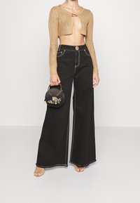 Donna che indossa un top corto in maglia dorato con maniche lunghe, jeans neri a vita alta e gamba larga, sandali dorati, e tiene in mano una borsa rotonda nera con design floreale dorato.