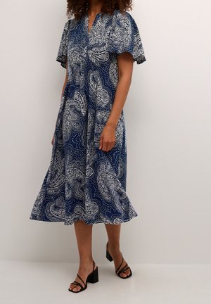 Abito blu navy con stampa paisley, maniche corte, scollatura a V, orlo morbido e delicati dettagli bianchi. Abbinato a sandali neri con tacco blocco e fasce.
