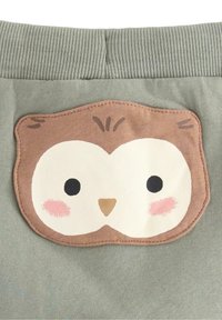 Zielona tkanina z teksturowanym pasem zawiera brązową łatkę w kształcie sowy z białą twarzą, czarnymi oczami i różowymi akcentami na policzkach.