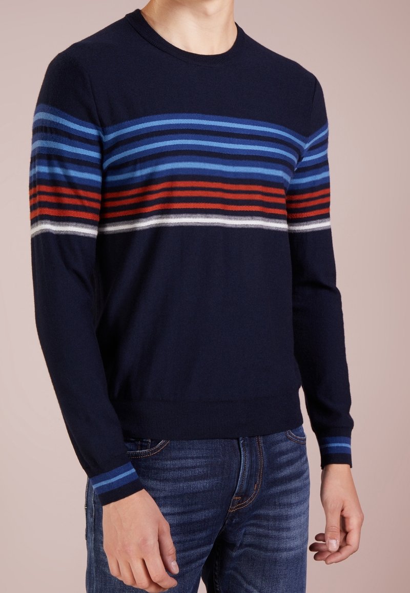 Homme portant un pull bleu marine avec des rayures horizontales bleues, rouges et blanches sur la poitrine et les manches, associé à un jean bleu.