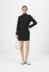 Robe noire à manches longues avec un col haut, au design ajusté. Le modèle tient un sac tissé gris clair et porte des chaussures gris clair à enfiler. Esthétique minimaliste.