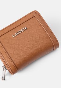 Brun strukturert lær lommebok med glidelås og sølv "LACOSTE" logo. Har synlige sømmer og en kompakt rektangulær form.