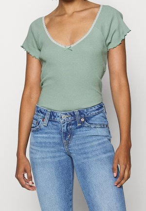 Jasnozielony prążkowany top z krótkimi falbankami na rękawach, z dekoltem w serek i małą kokardką, zestawiony z niebieskimi dżinsami.
