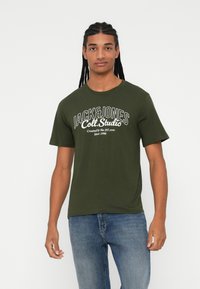 Olive roheline puuvillane T-särk, millel on ümar kaelus, lühikesed varrukad ja valge trükitud tekst "JACK&JONES Coll. Studio" ees.