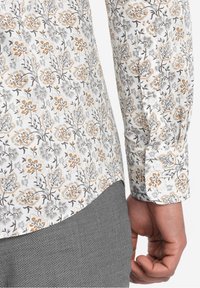 Langarmshirt van witte stof met bloemenprint in grijs en goud; knoopmanchetten; smalle geruite patroon eronder.