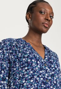 Blusa de estampado floral em azul marinho com flores brancas e coloridas, fecho de botão e ombros franzidos. Tecido suave e design casual.