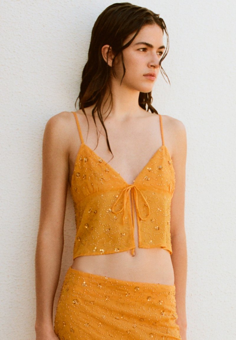 Jeune femme aux cheveux mouillés portant un crop top orange scintillant à fines bretelles et une jupe assortie, debout contre un mur blanc.