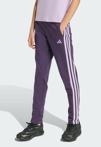 Fialové športové nohavice so bočnými vreckami, s tromi svetlofialovými pruhmi a logom Adidas na ľavom stehne. V kombinácii s čiernymi teniskami.