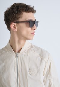 Beige bomberjack met een gladde textuur, voorrits en subtiel berenlogo; gecombineerd met zwarte rechthoekige zonnebril.