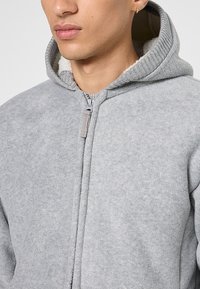 Młoda osoba ubrana w jasnoszary polarowy sweatshirt zapinany na zamek, z miękką białą podszewką widoczną w kapturze, z bliska ukazująca szyję i dolną część twarzy.