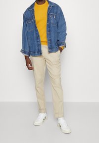 Jeansjacke in Blau, gelber Rippstrickpullover darunter, beigefarbene Hose und weiße Turnschuhe mit grünen Akzenten. Lässige Passform, klassisches Design.