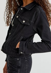 Mujer vestida con una chaqueta de denim negra con botones de metal, con las manos en los bolsillos, combinada con pantalones de denim negros, frente a un fondo claro y liso.