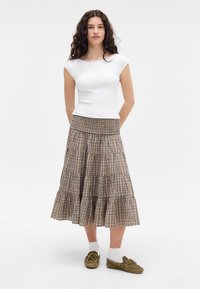 Jupe midi tiered à carreaux bruns avec une taille smockée, associée à un t-shirt blanc ajusté et des mocassins vert olive.