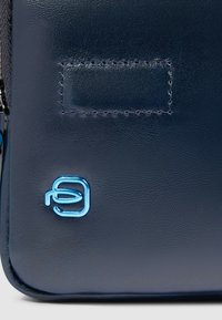 Marineblaue Ledertasche mit glatter Textur, die ein auffälliges blaues Logo-Akzent und einen rechteckigen, gesteppten Aufnäher zeigt.