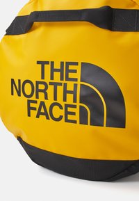 Round duffel bag in bright yellow material with a black textured bottom, featuring large black logo text, and a sturdy black strap.