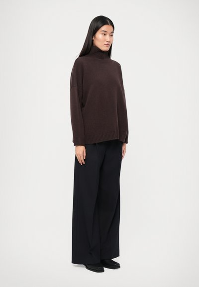 Pull à col roulé marron avec des poignets côtelés, associé à un pantalon noir à jambes larges et des chaussures noires, porté par une personne se tenant devant un fond blanc.
