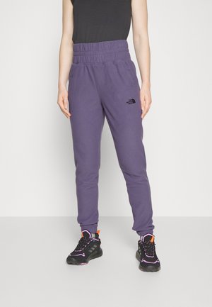 Persoon draagt paarse trainingsbroek met hoge taille en The North Face-logo, en zwarte sneakers met oranje en roze accenten, staand op een grijze vloer.