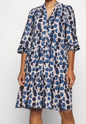 Frau trägt ein knielanges Kleid mit dreiviertel Puffärmeln, das ein blau-schwarzes Tupfenmuster auf hellem Hintergrund zeigt.