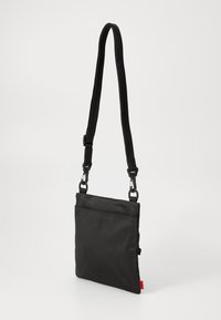 Sac bandoulière en tissu noir avec bandoulière réglable, présentant une forme carrée simple, une texture lisse et un détail de marque rouge.