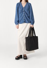 Femme portant un chemisier bleu boutonné et une longue jupe blanche, tenant un grand sac fourre-tout noir avec des sandales à plateforme noires hautes.