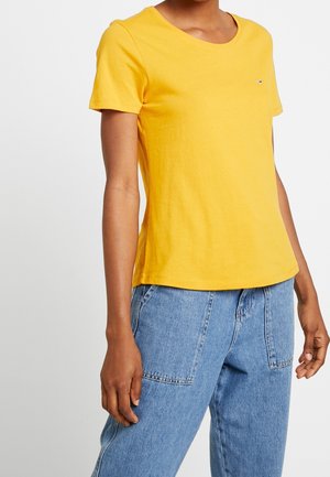 Camiseta de algodón amarilla de manga corta con cuello redondo y un pequeño logo de la marca en el lado izquierdo del pecho, combinada con jeans azul claro.