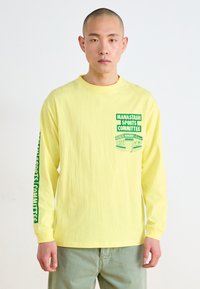 Camiseta de manga larga amarilla con texto y gráficos en verde, con un cuello redondo y un ajuste relajado. El texto en la manga y el pecho añade detalle.