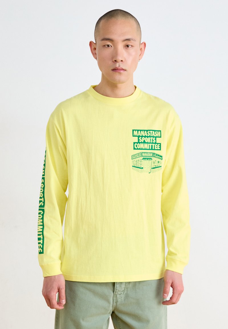 Camiseta de manga larga amarilla con texto y gráficos en verde, con un cuello redondo y un ajuste relajado. El texto en la manga y el pecho añade detalle.
