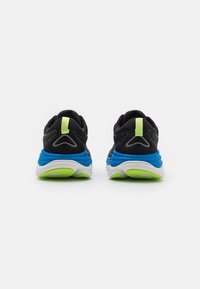 Des chaussures de sport noires avec une semelle intermédiaire bleue et une semelle extérieure verte. La vue arrière montre une languette de tirage verte vive et des détails texturés.