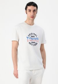 White cotton t-shirt featuring graphic print: "ORIGINAL 1985 TOMMY JEANS NEW YORK" in blue, red, and white colors with a relaxed fit.