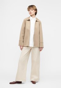 Νέος άνθρωπος όρθιος, φοράει μπεζ παντελόνι φαρδιάς γραμμής, άσπρο πουκάμισο με κουμπιά, ανοιχτό καφέ μπουφάν με φερμουάρ και σκούρα καφέ παπούτσια σε απλό φόντο.