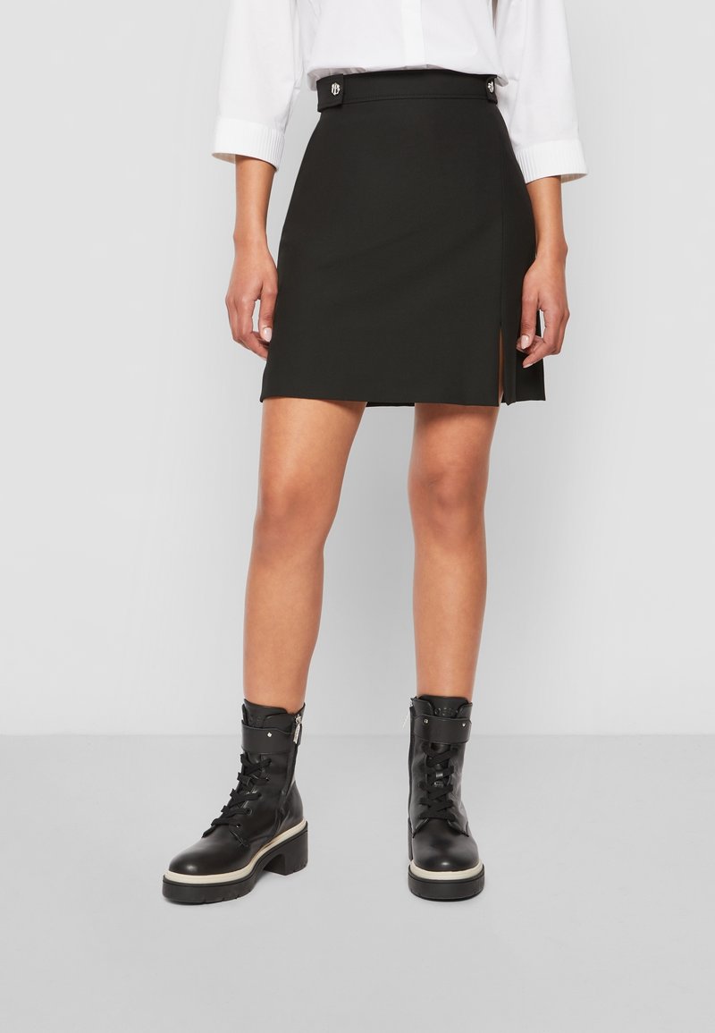 Černá přiléhavá mini sukně s bočním rozparkem, vyrobená z hladkého materiálu. Kombinována s černými šněrovacími botami s masivní podrážkou.
